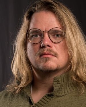 Portrait of someone with round glasses and blonde hair wearing a green sweater.