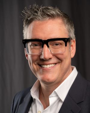 Professional headshot of someone in glasses wearing a dark suit jacket.