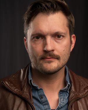 Portrait of someone in a brown leather jacket and denim shirt.