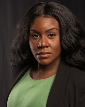 Professional portrait of someone in a black blazer and green top against a dark background.