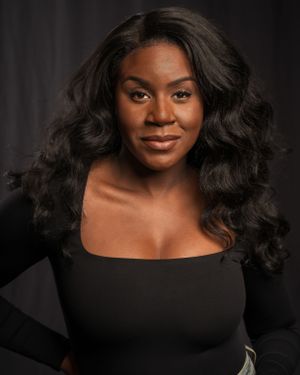 Professional studio portrait of a woman with voluminous dark curls wearing a black top against a dark background.