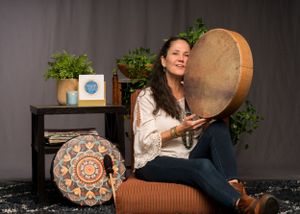 Musician seated with traditional frame drums and potted plants in a studio setting.