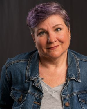 Casual portrait of a person with purple hair wearing a denim jacket against a dark background.