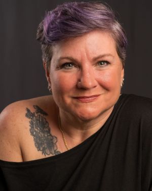 Studio portrait of a person with purple-tinted short hair wearing a black top showing a shoulder tattoo.