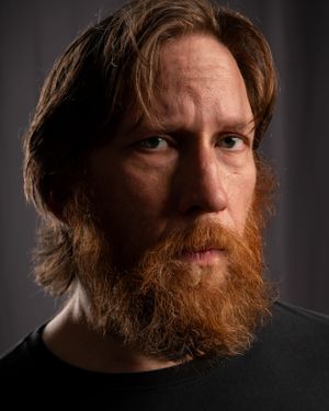 Dramatic side-lit portrait of a person with auburn beard and hair against dark background.