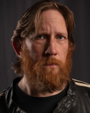 Close-up portrait of a person with long red beard wearing black against dark backdrop.