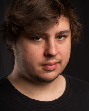 Studio headshot of a young person with tousled brown hair wearing a black t-shirt.