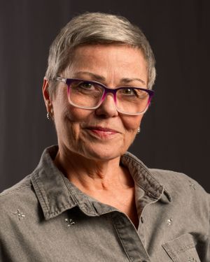 Professional headshot of mature person with short gray hair wearing glasses and denim shirt.