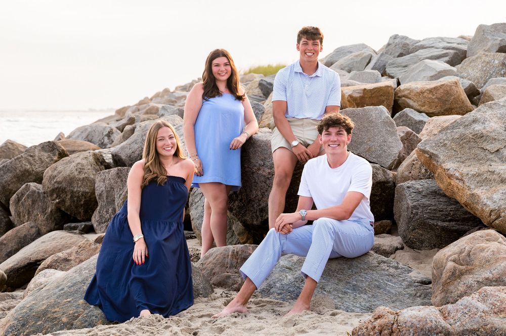 Four teenagers pose casually on rocky beach in coordinating blue and white outfits for family portrait.