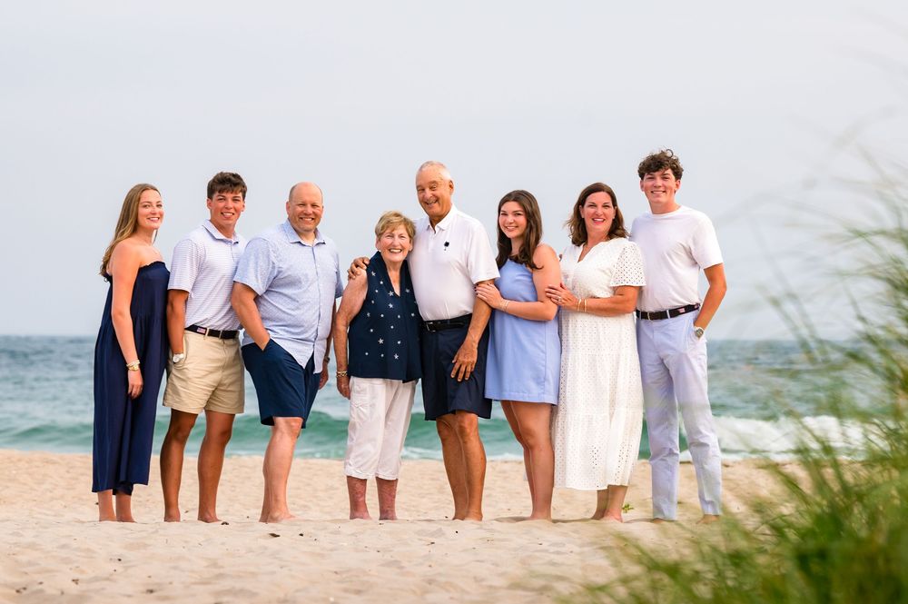 Extended family of eight poses together smiling on sandy beach with turquoise ocean in background.
