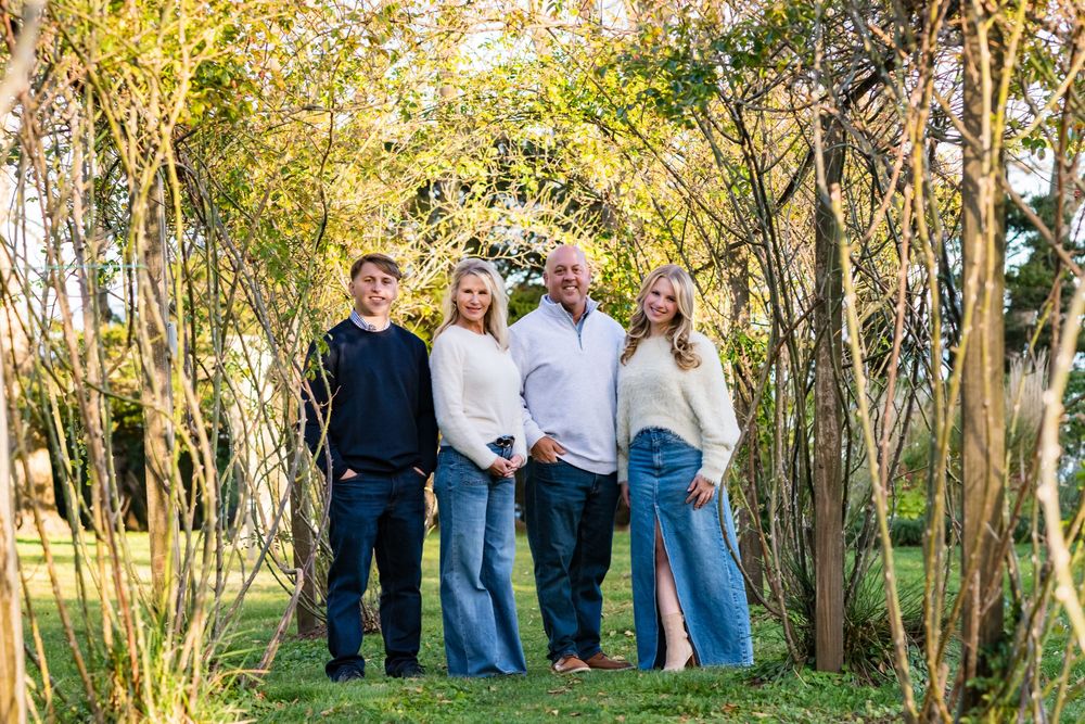 Family of four smiling together under a natural archway of trees in a garden.