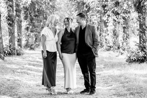 Two women and a man laugh together while walking on a sunlit forest path in black and white.