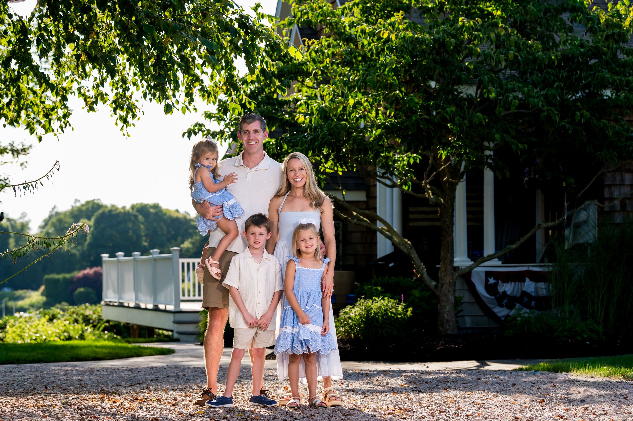 Happy family of five posing outdoors near a white bridge surrounded by lush green trees on a sunny day.