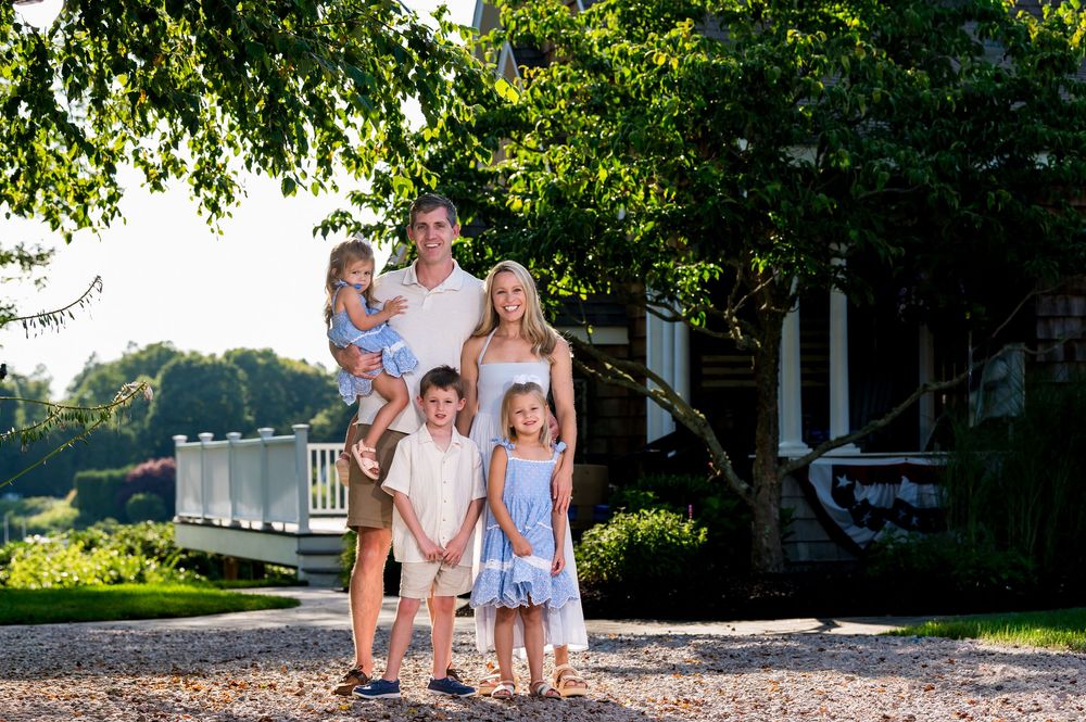 Happy family of five posing outdoors near a white bridge surrounded by lush green trees on a sunny day.
