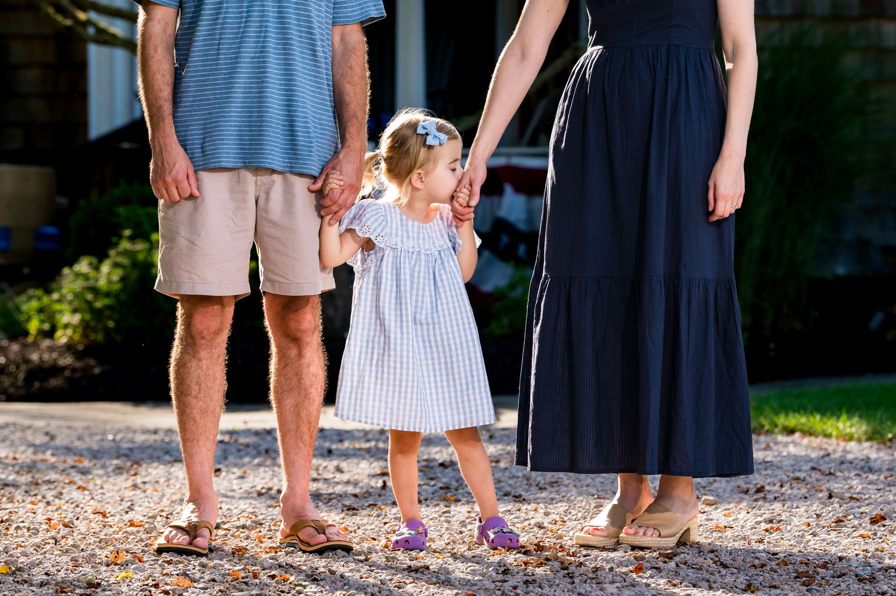 Young toddler girl in striped dress holds hands with parents standing on gravel path outdoors.