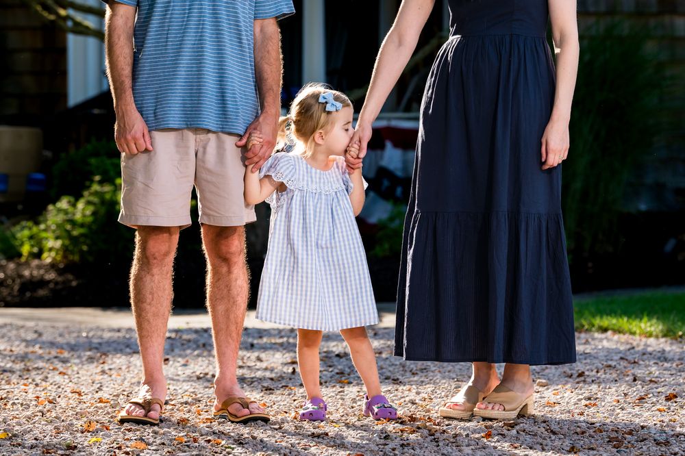 Young toddler girl in striped dress holds hands with parents standing on gravel path outdoors.