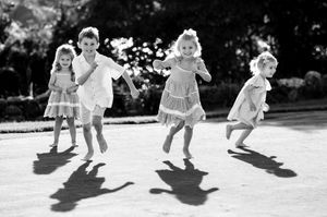 Four young children jump and run playfully outdoors casting long shadows in black and white photo.
