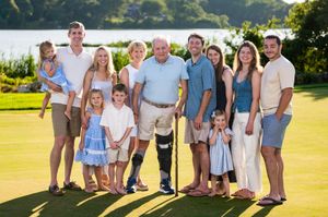 Large multigenerational family poses together smiling on green golf course near scenic waterfront at sunset.