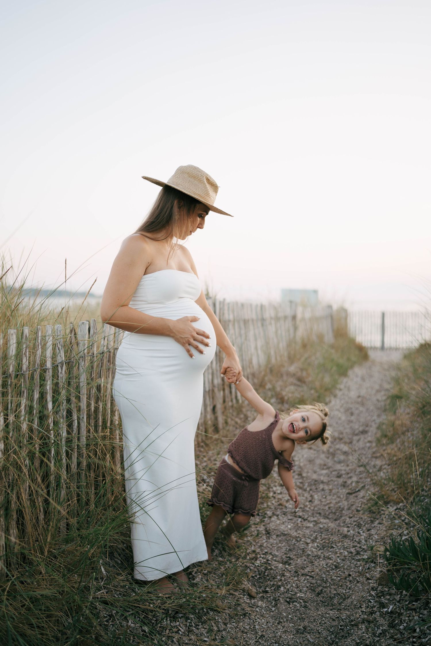 Une séance photo grossesse maternité montrant une femme enceinte en robe blanche posant au coucher du soleil sur la plage.