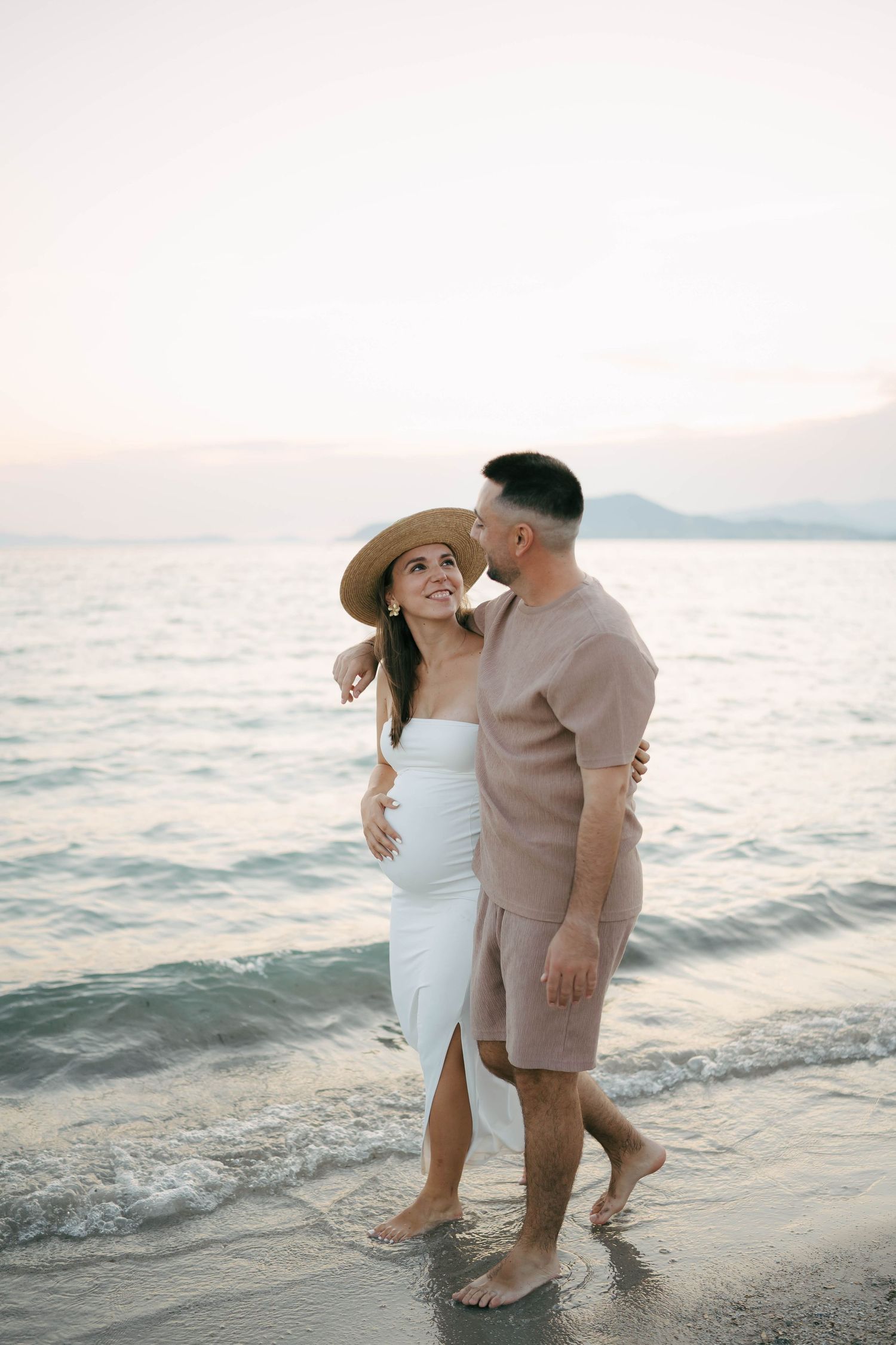 Romantic beach photoshoot with couple in light-colored attire dancing by the ocean during sunset.
