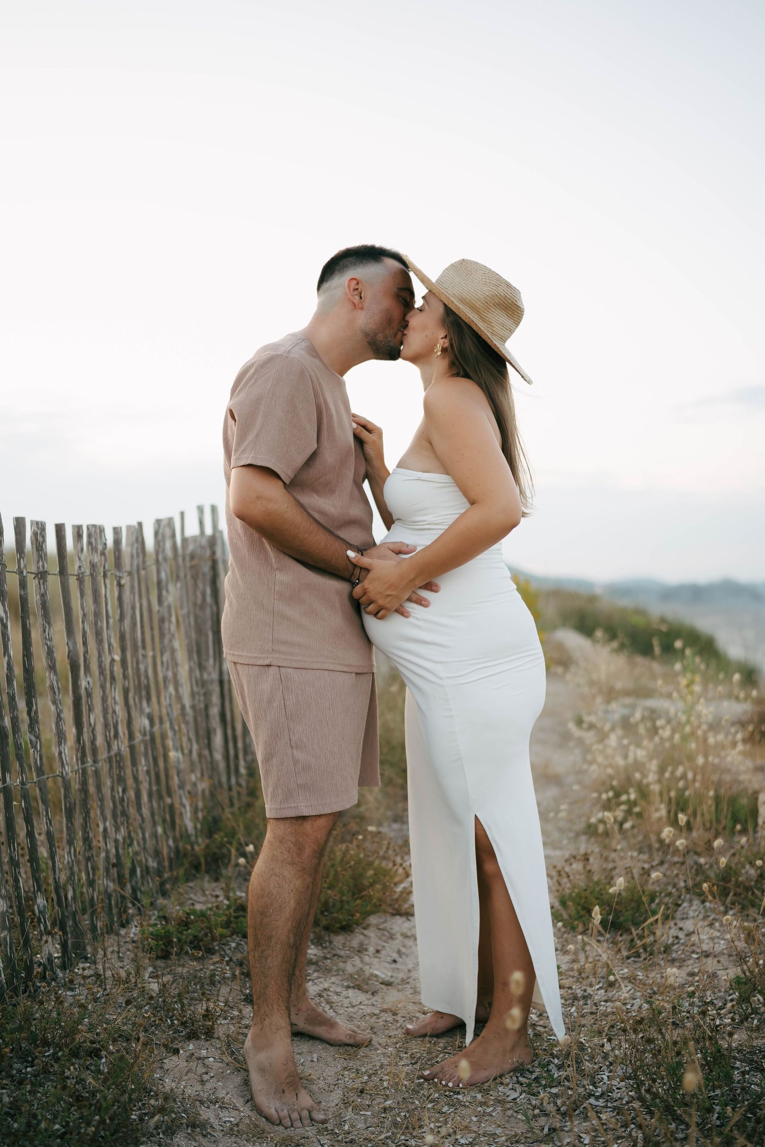 A romantic couple embraces on a hilltop at sunset beside a rustic wooden fence, she in white and he in tan.