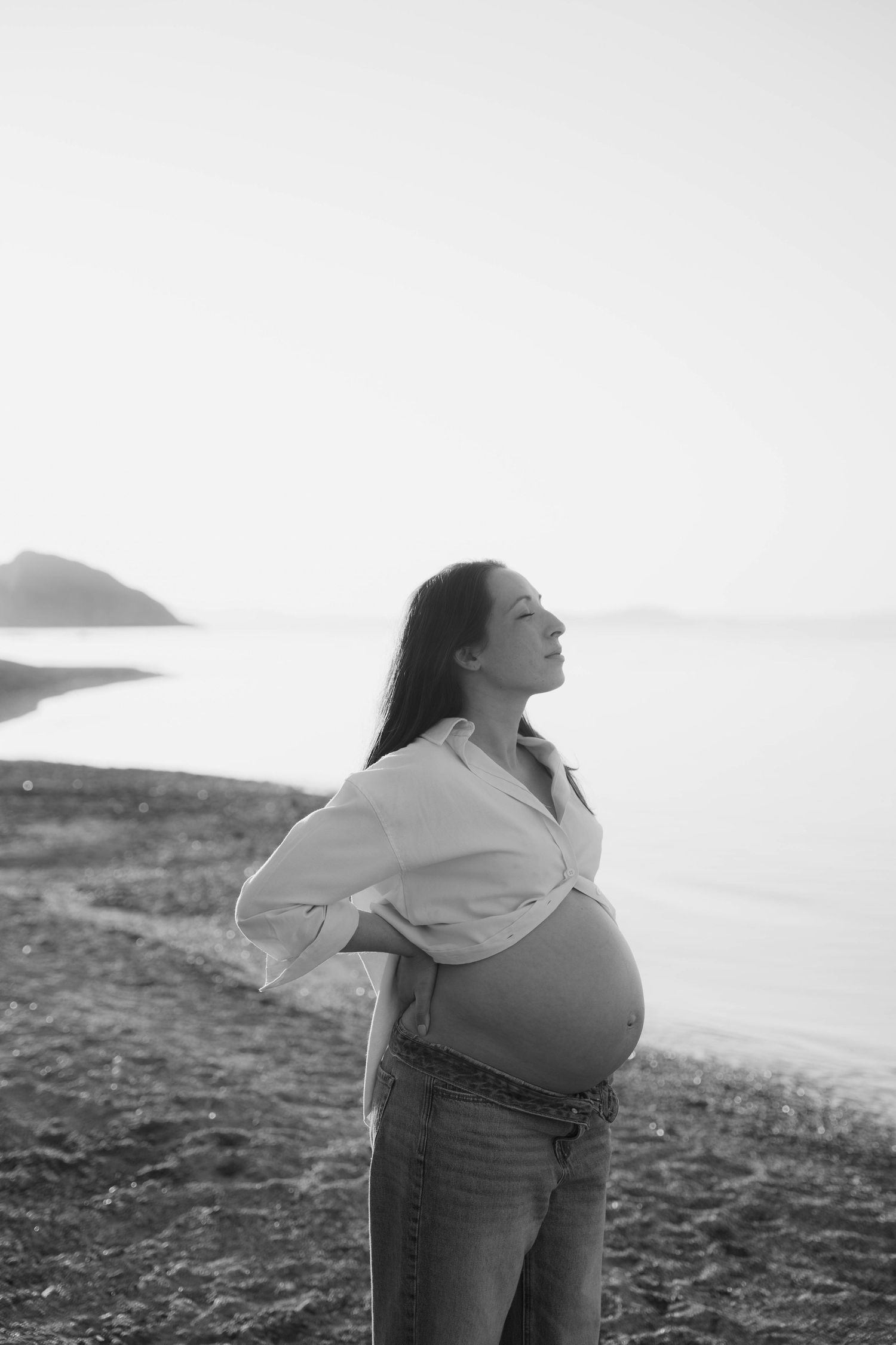 A person in jeans leans back with arms outstretched against a coastal landscape in black and white photography.