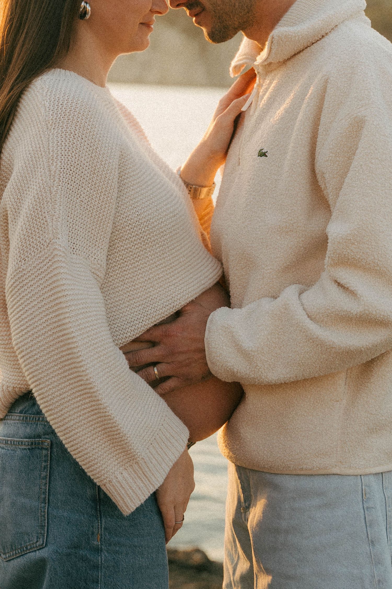 A romantic couple in matching cream sweaters and light denim embrace in warm sunlight.