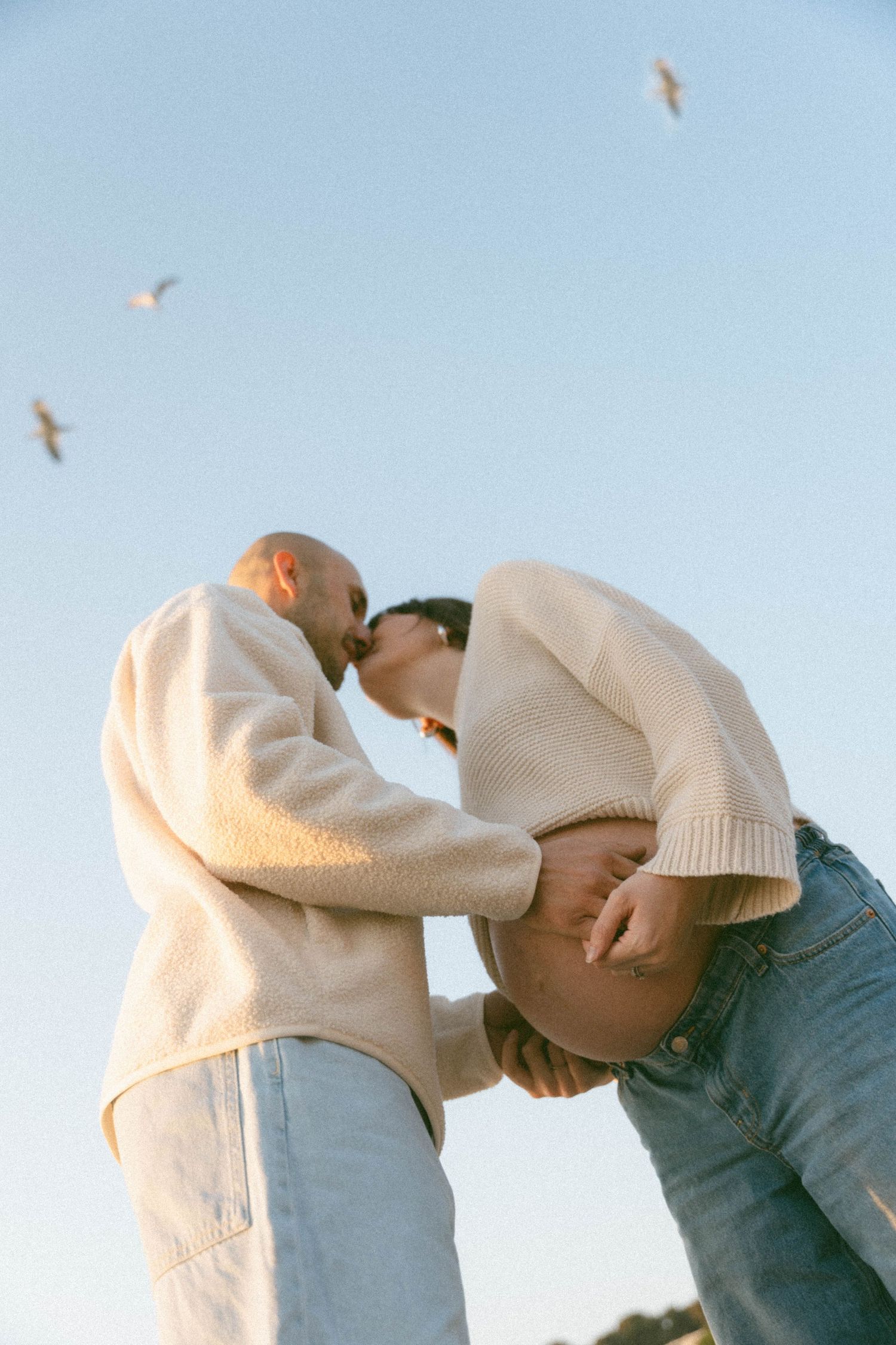 Two people in white and blue clothing lean in close to each other against a bright sky with birds flying overhead.