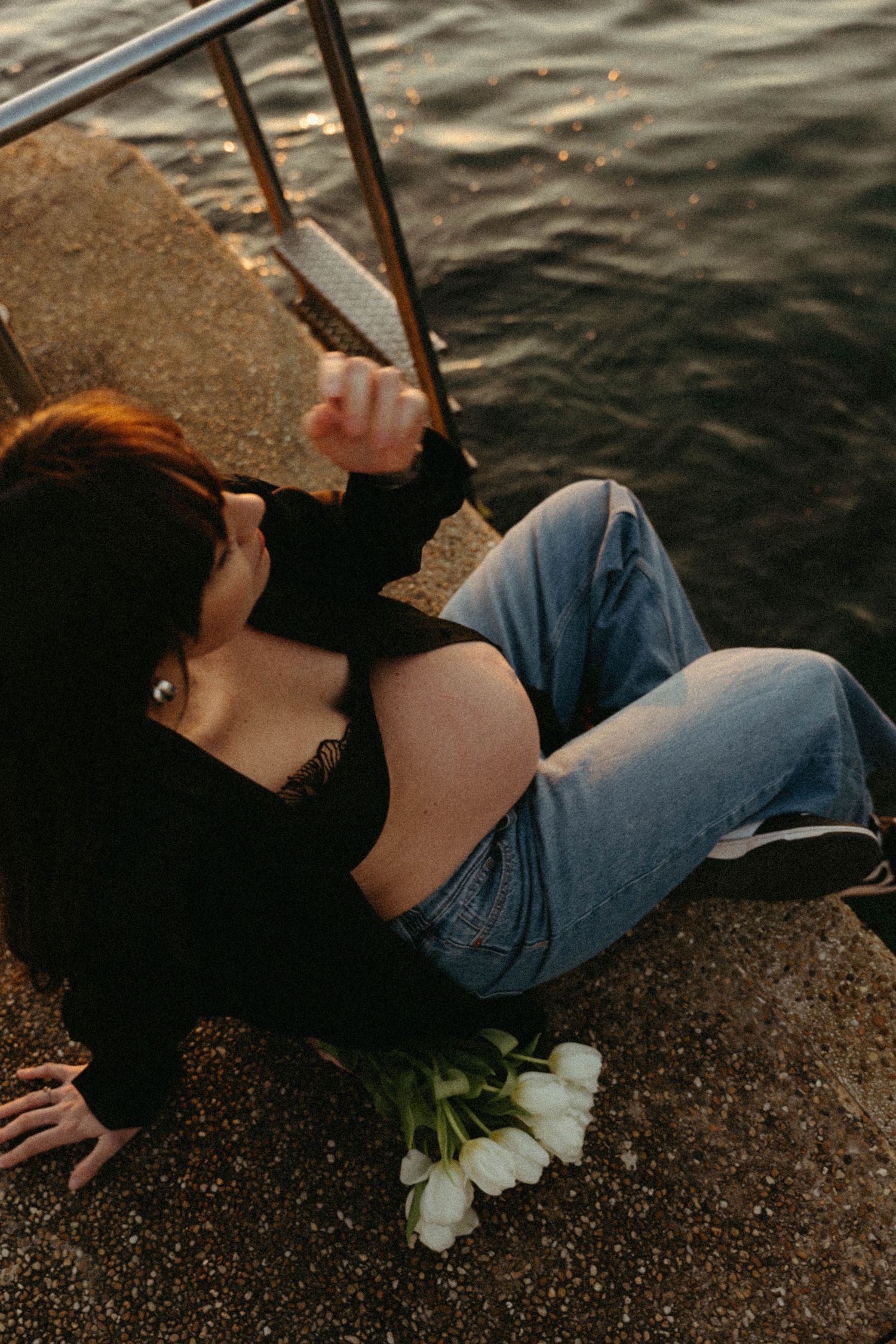 A person lies on a stone surface near water while white flowers rest beside them in a moody outdoor setting.