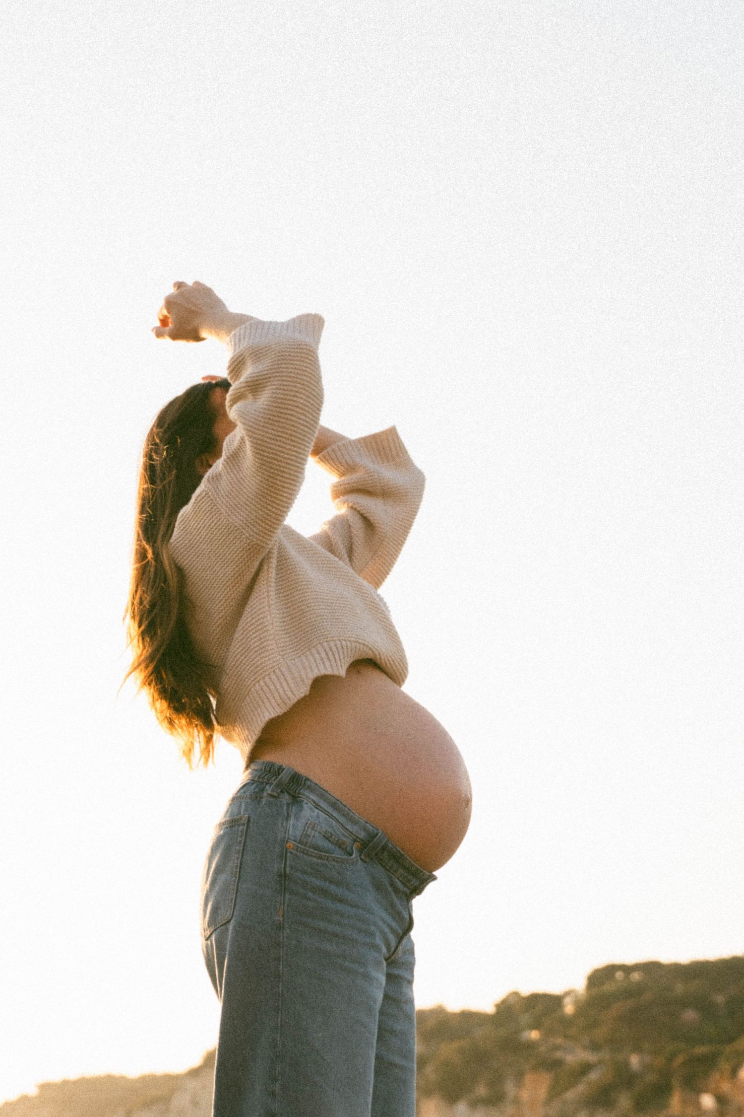 A pregnant person in jeans stands against a sunset sky, photographed in a series of three similar poses.