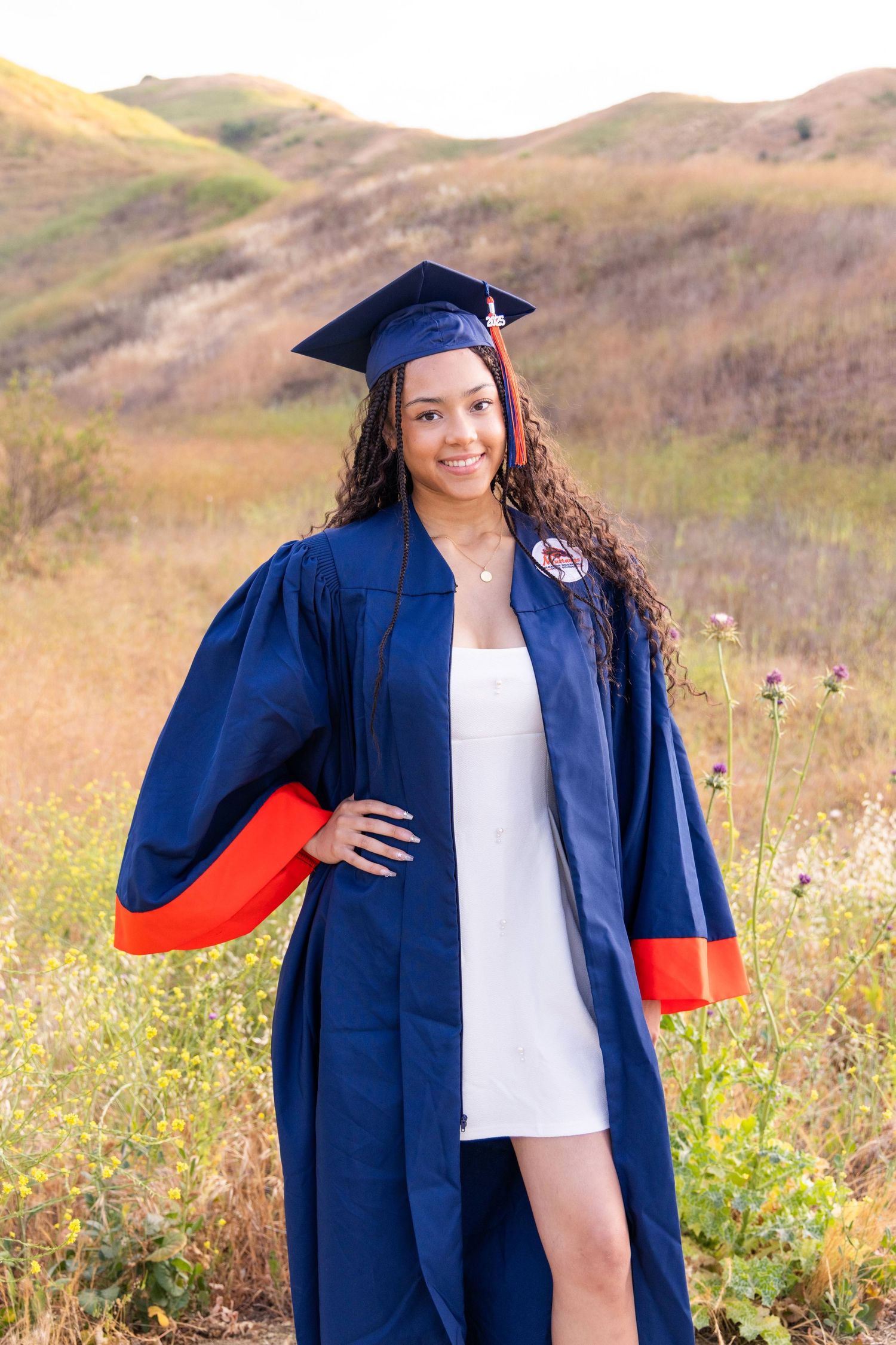 Smiling graduate in navy blue cap and gown posing in a field with rolling hills in the background.