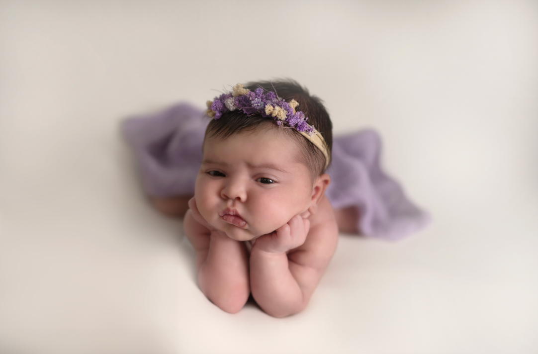 Newborn baby girl posed froggy style on white backdrop wearing purple floral headband and wrapped in lavender fabric.Embracing Life’s Beauty
