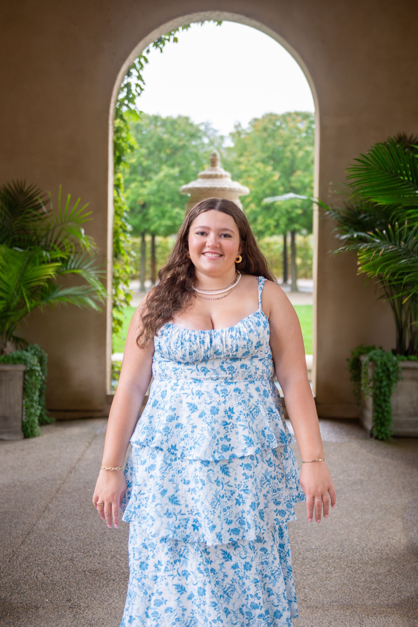 Senior portrait in blue floral dress posing under arched walkway with plants.
