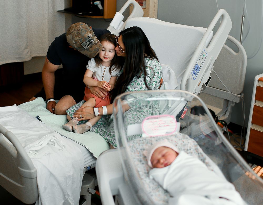 Mom, dad and big sister sitting in hospital bed while newborn lays in her bed for fresh 48 session