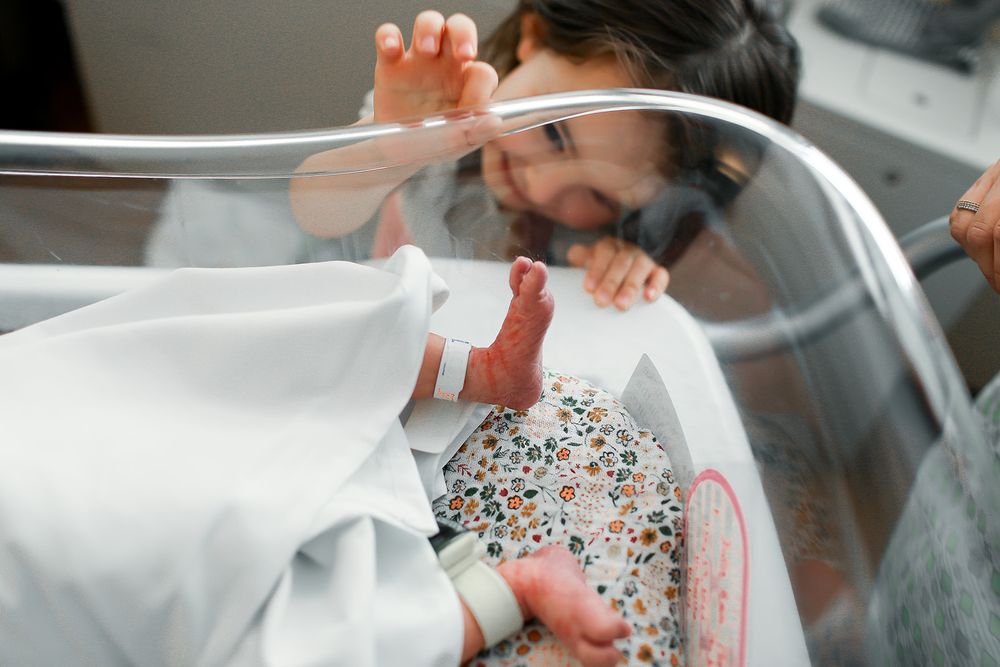 Big sister peeking over hospitall crib looking at baby sister
