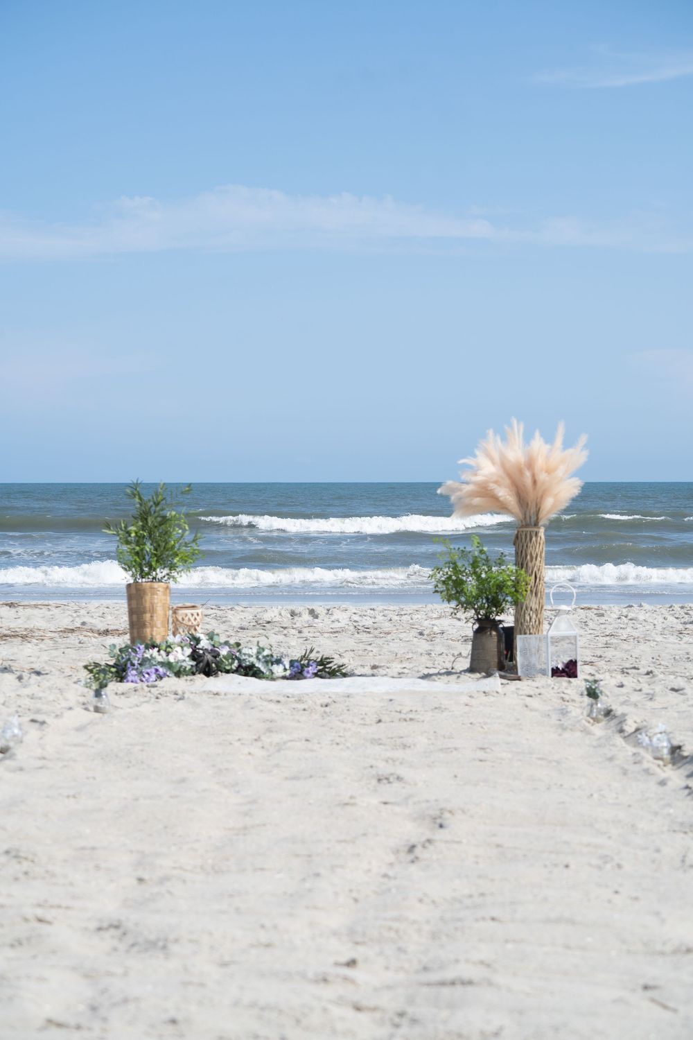 Beach wedding decorations with vases and flowers line the sandy shore against a blue ocean backdrop.