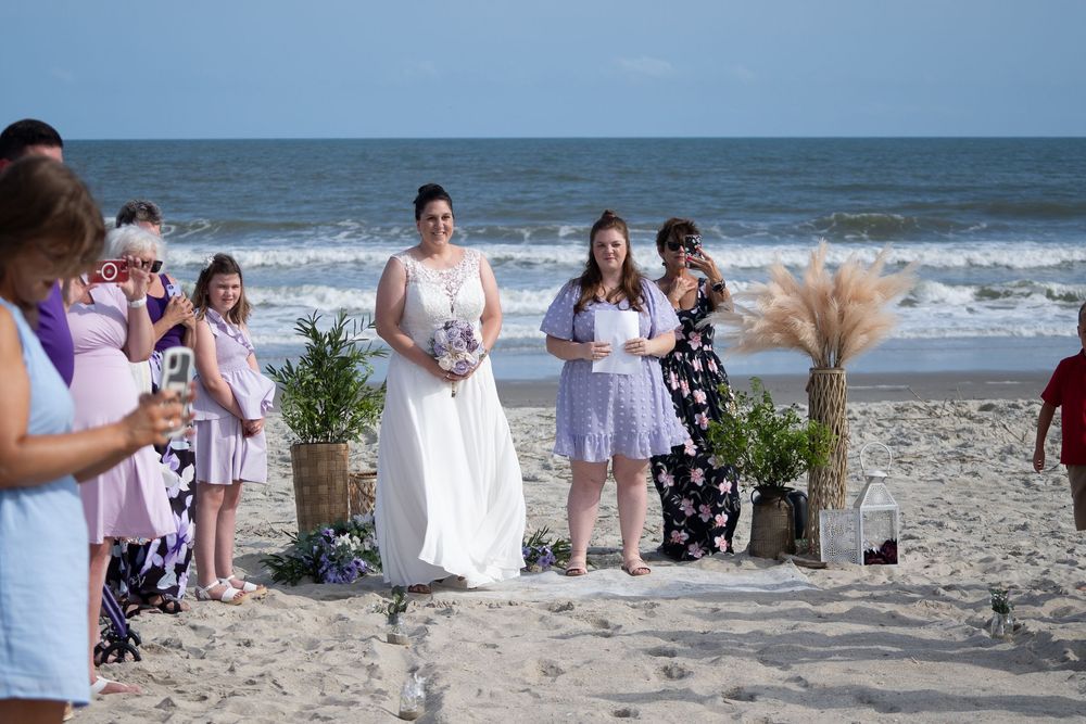 A group of wedding guests gather for a daytime ceremony on a scenic ocean beach with waves visible in the background.