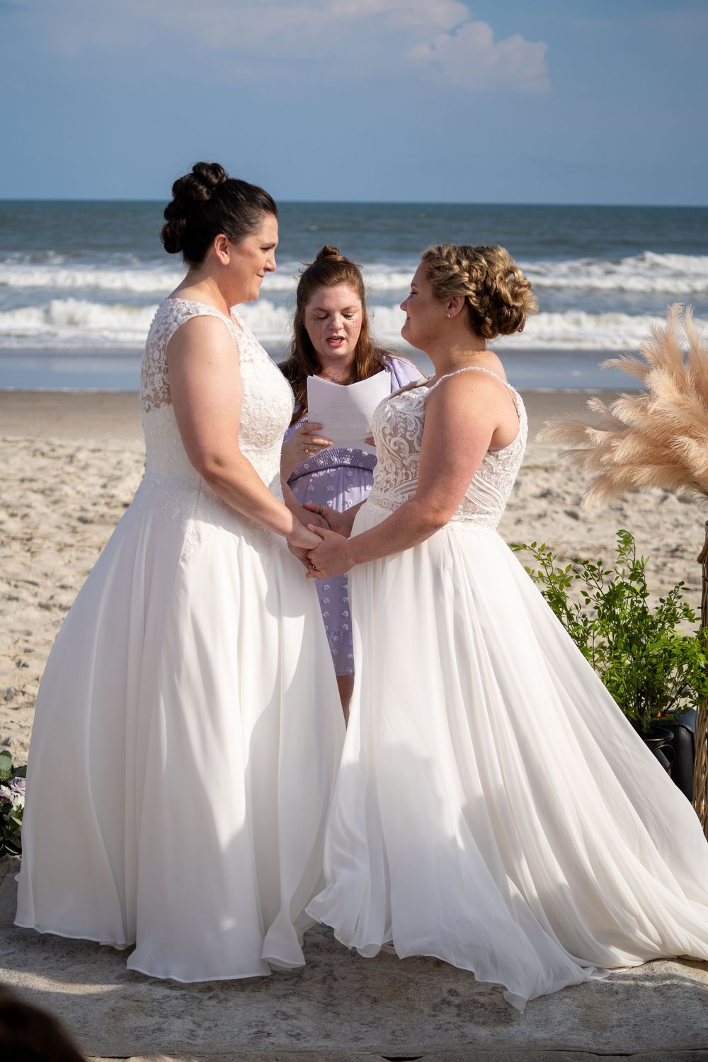 White-gowned couple shares tender moments with officiant during intimate beach wedding ceremony on sunny day.