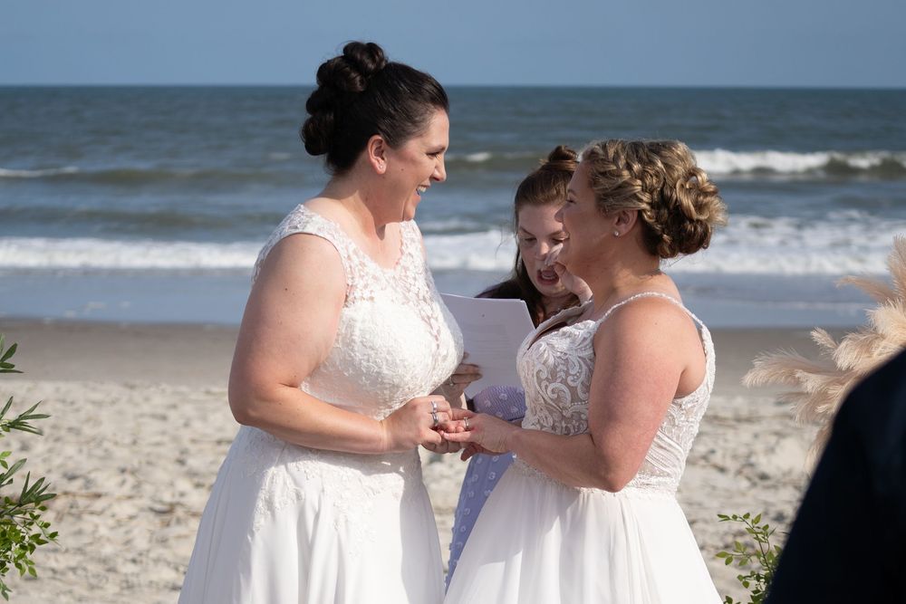 Two people in white lace wedding dresses share a joyful moment together on a sunny beach by the ocean.