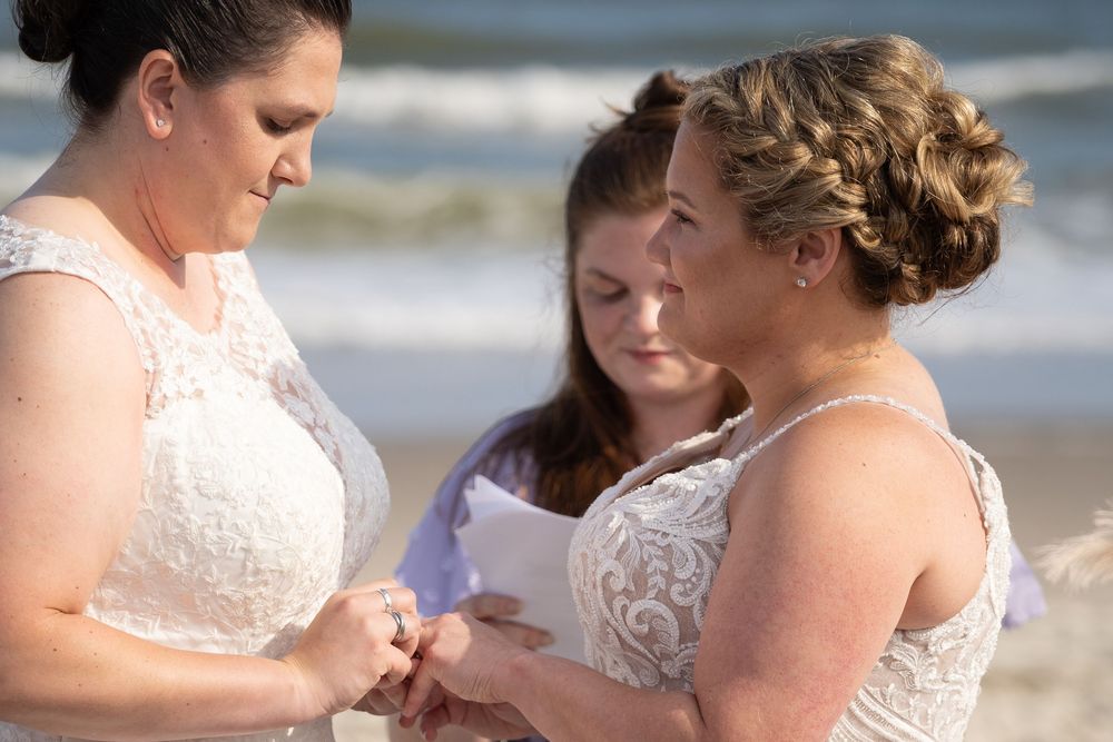 Two people share an intimate moment during a beach wedding ceremony with waves and sand in the background.