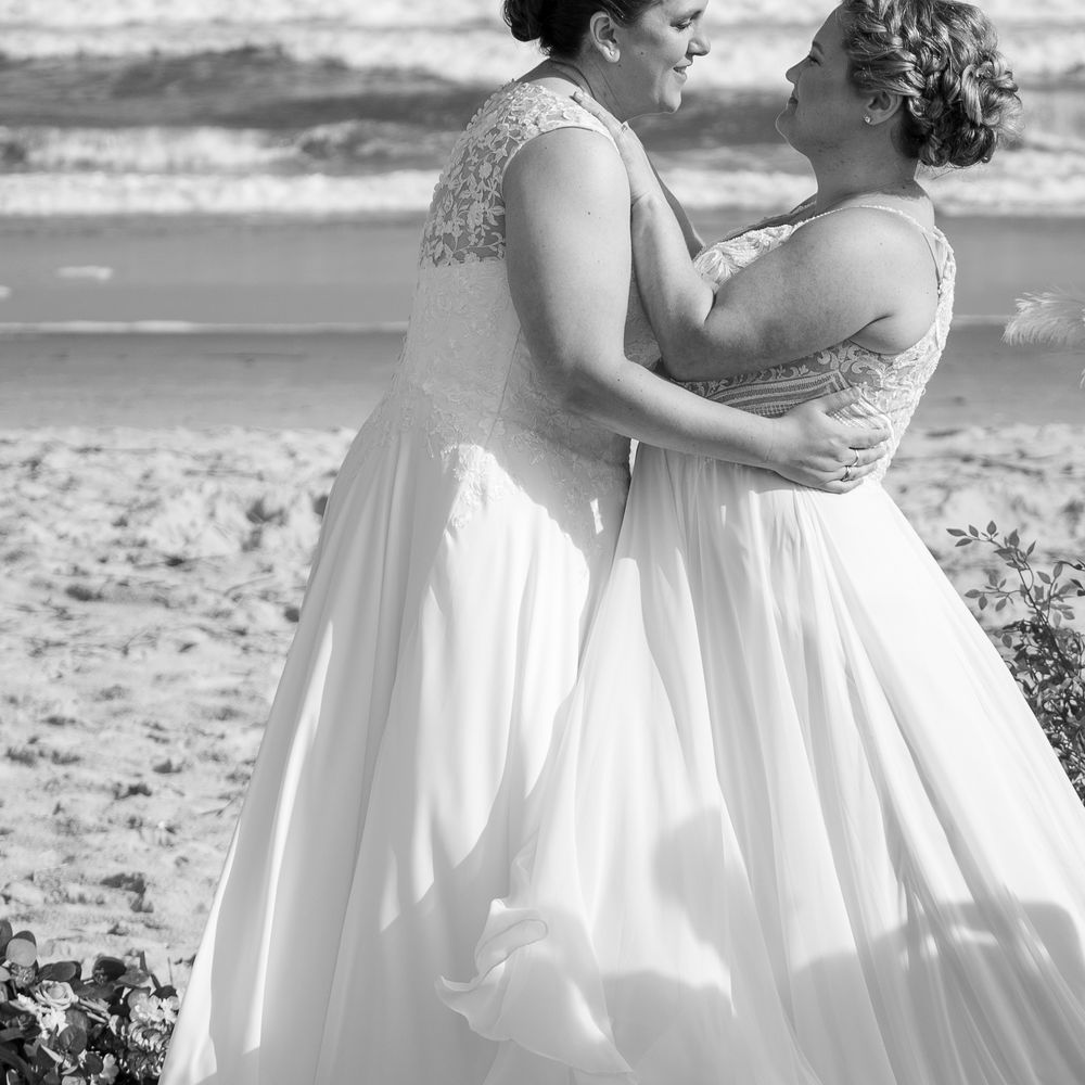 Two people in white dresses share a sweet intimate embrace on a sunlit beach during a romantic moment together.