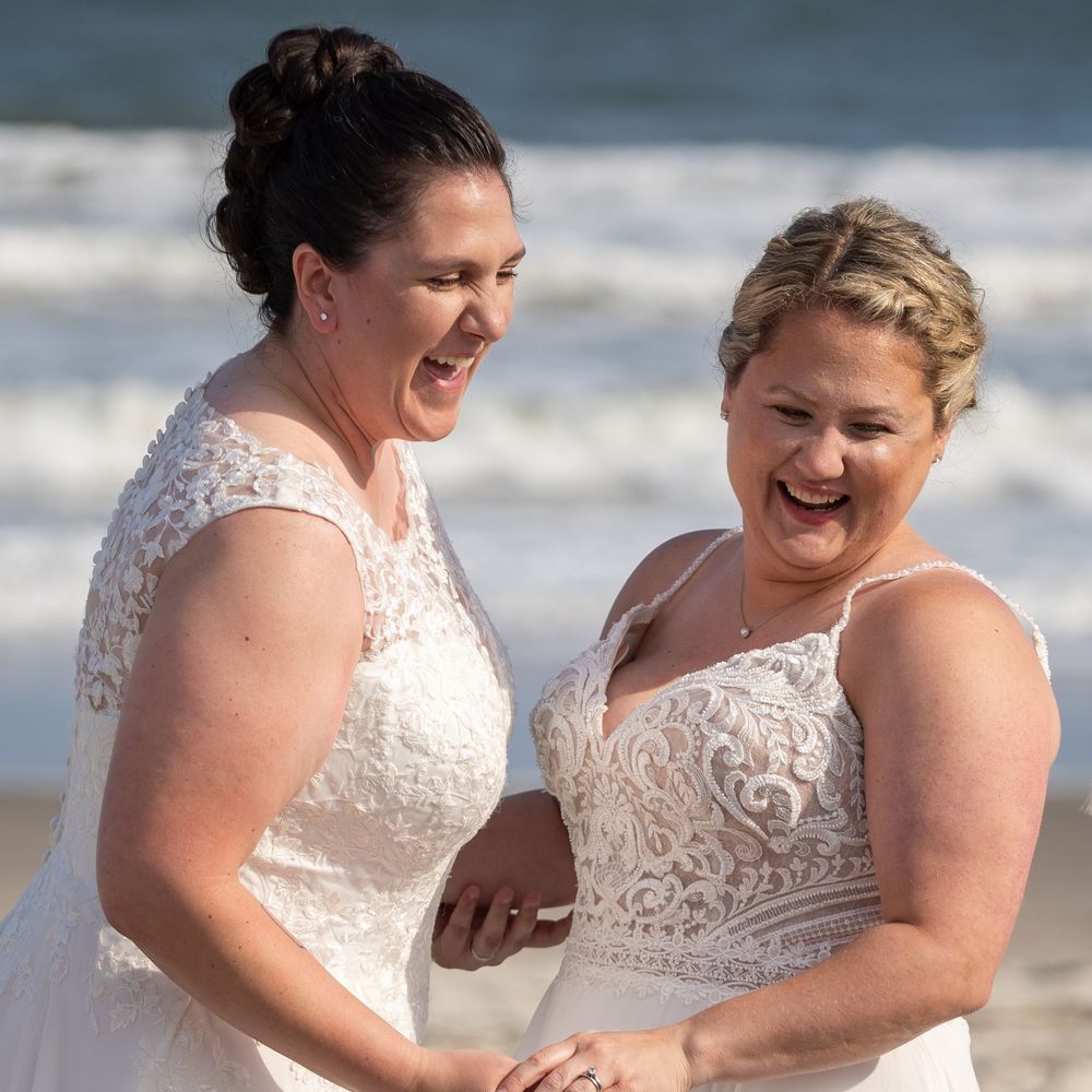Two people in white lace dresses holding hands and laughing together on a sunny beach.