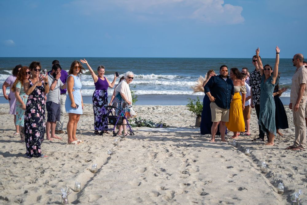 A large group celebrates during a beach wedding ceremony as waves crash on the shoreline in the background.