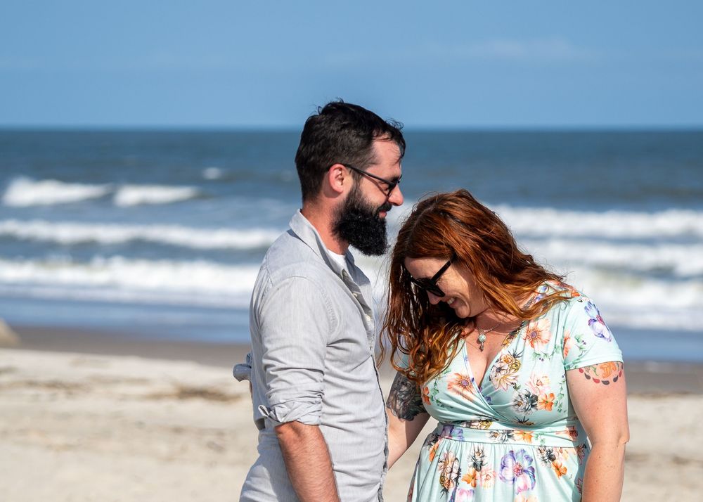 A couple walks together holding hands along a peaceful beach with ocean waves and blue skies in the background.
