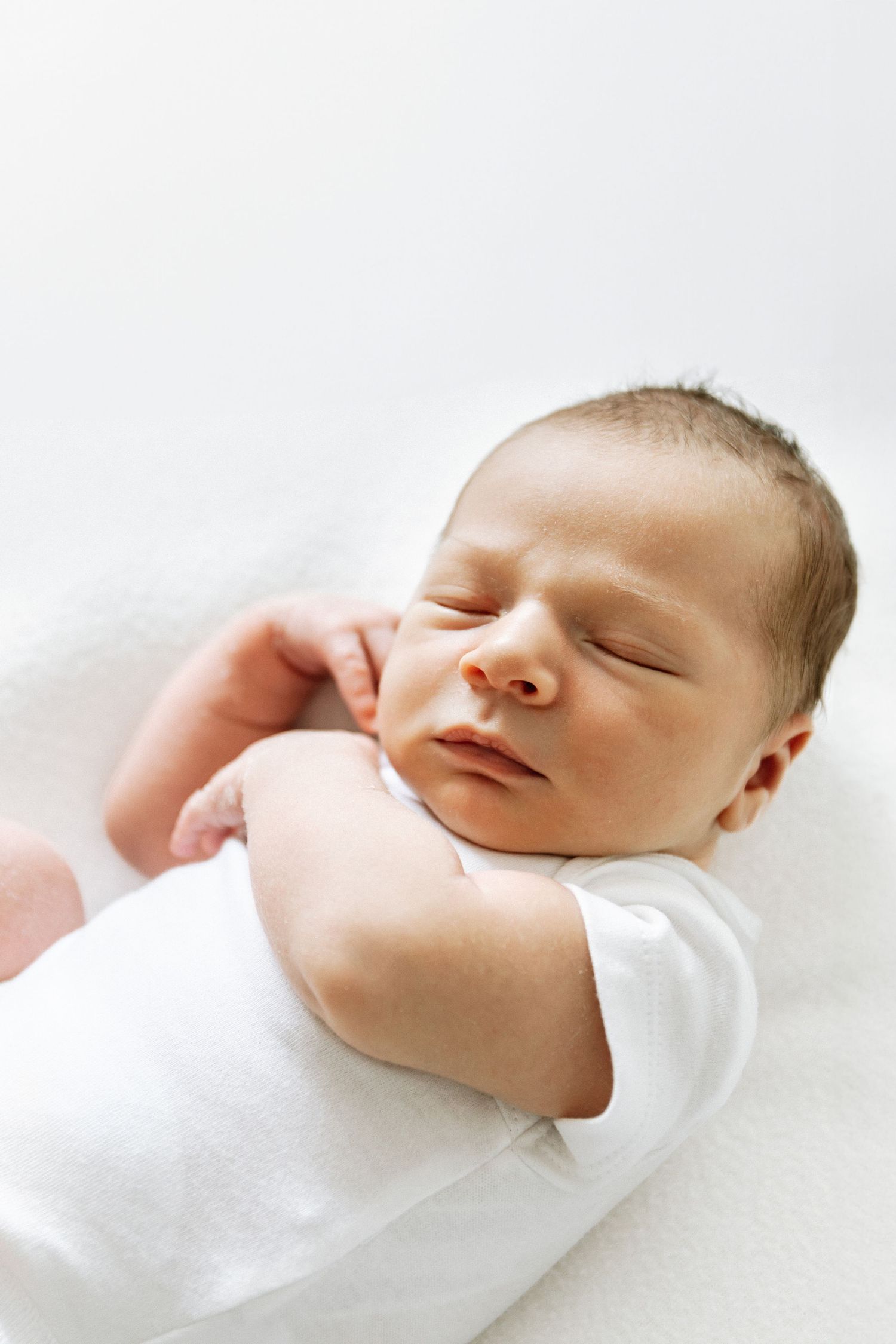 A sleeping newborn baby wearing white lies peacefully with closed eyes against a white background during newborn shoot in Nottingham