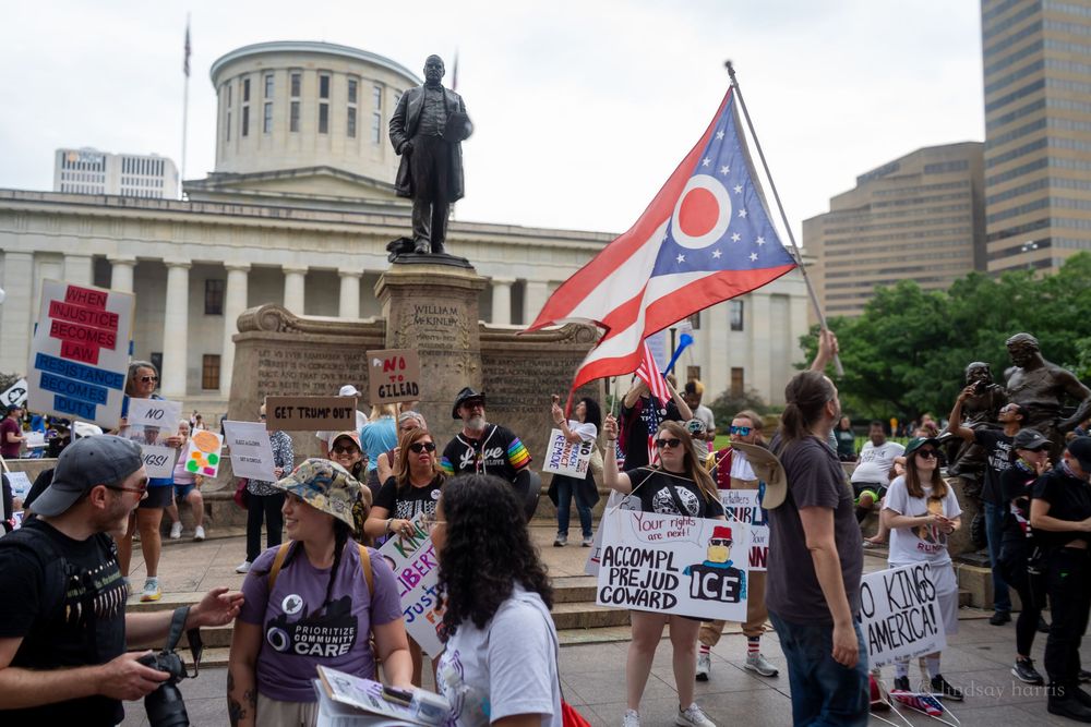 Group of demonstrators gather at the Ohio Statehouse in Columbus during a protest rally with flags and signs.