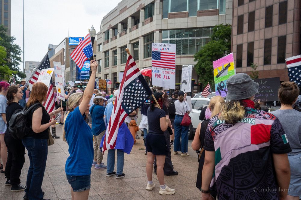 Crowd of protesters waving American flags during a street demonstration amidst urban buildings.