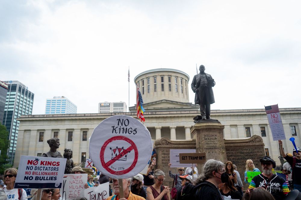 Panoramic view of protestors holding signs at a rally in front of a government building with domed architecture.