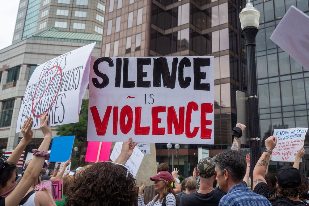 Crowd of protesters hold signs at a rally against violence and silence with buildings visible in background.