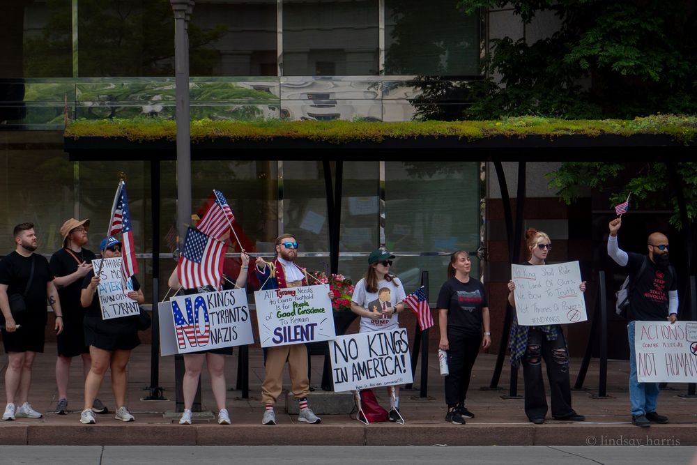 Protesters line a street holding American flags and handwritten signs during a public demonstration.
